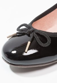 Ballerine en cuir verni noir avec un nœud noir tressé et des extrémités dorées sur une doublure intérieure rose clair.