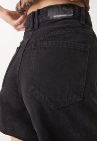 Mustat farkkushortsit korkealla vyötäröllä, jossa on nahkainen tagi nimeltä "STRADIVARIUS" ja näkyvät takataskut ompeluyksityiskohdilla.