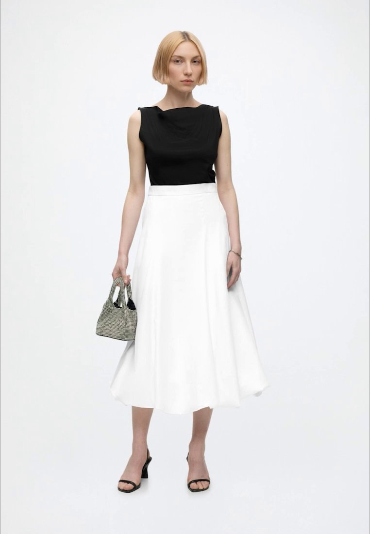 Femme portant un haut noir sans manches et une jupe midi blanche, tenant un petit sac à main à carreaux, portant des talons ouverts noirs, debout sur fond blanc.