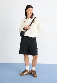 Weißes Langarm-Polo mit bordeauxfarbenem Kragen und gesticktem Logo, kombiniert mit schwarzen Shorts und braunen Sneakern mit weißen Streifen.