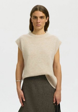 Vrouw draagt een mouwloos beige gebreid topje en een donkergrijze rok, staand tegen een effen achtergrond en houdt een zwarte tas vast.