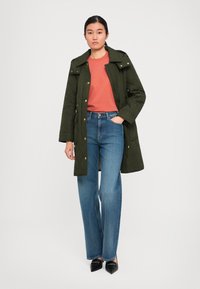 Femeie îmbrăcată într-o haină cu quilted de culoare verde măslin, cămașă de culoare ruginie, blugi albaștri și pantofi cu vârf ascuțit negri, stând cu o mână în buzunar.