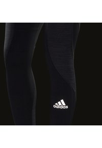 Leggings sportivi neri con un motivo testurizzato, caratterizzati da un logo Adidas bianco riflettente sulla gamba inferiore. Materiale liscio e design aderente.