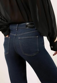 Jeans din denim albastru închis, cu o formă ajustată, având două buzunare posteriori, cusături în contrast și o etichetă de talie din piele neagră.