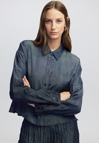 Chemise bleu foncé à boutons avec un col structuré, un tissu texturé et des manches longues, dotée de plusieurs boutons et d'un design court.