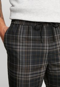Primo piano di pantaloni a quadri neri e marroni con coulisse indossati con una maglietta grigia chiara, che mostrano una mano tatuata appoggiata nella tasca.