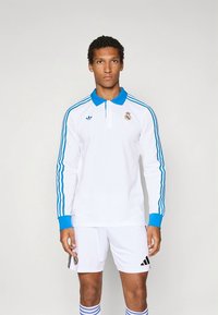 Λευκό πόλο με μακριά μανίκια με μπλε λεπτομέρειες και ρίγες Adidas, με λογότυπο της Ρεάλ Μαδρίτης στο στήθος και μπλε γιακά.