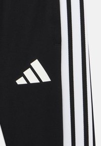Schwarze Sporthose mit drei weißen Streifen an den Seiten und einem weißen Logo. Der Stoff hat eine glatte, matte Textur.