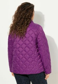 Giacca trapuntata viola con un motivo a diamante, caratterizzata da un colletto alto e una texture liscia e lucida. La vista posteriore mostra le maniche lunghe e l'orlo.