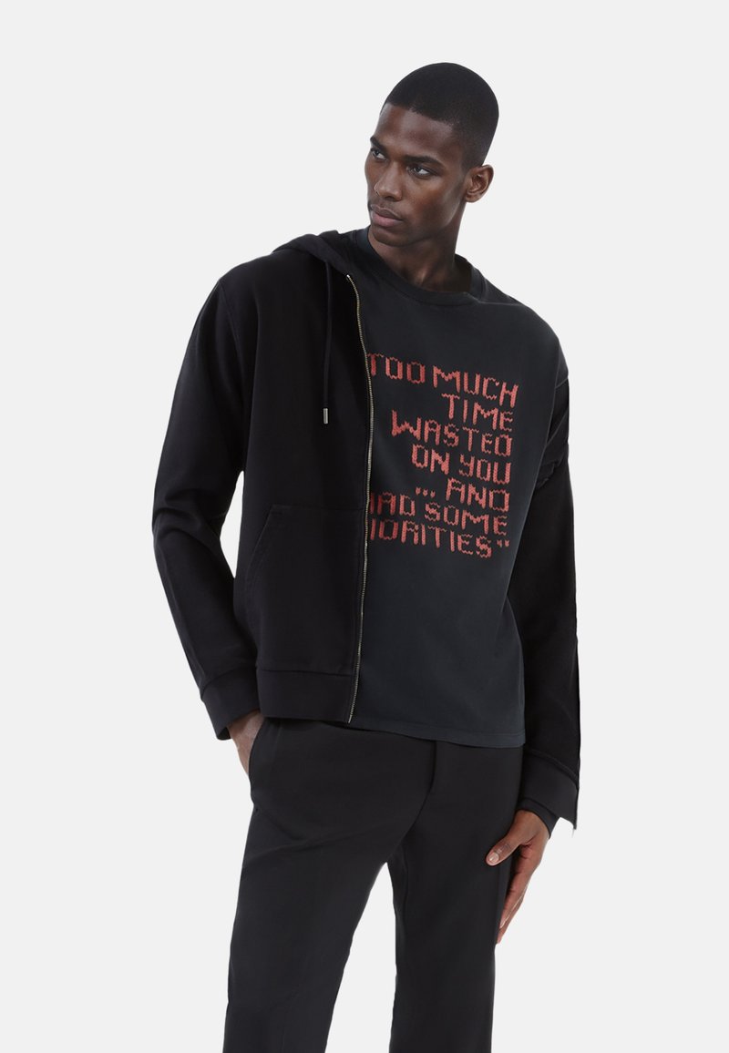 Sort zip hoodie med kængurulomme, lagd over en sort sweatshirts med rød tekst. Glat tekstur, afslappet pasform, minimalistisk design.