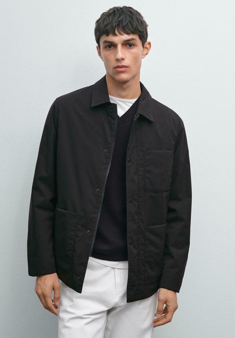 Young man wearing a black buttoned jacket over a black sweater and white pants, standing against a light gray background.