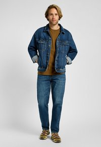 Denim-takki rentoa istuvuutta, vaaleansininen väri, kaksi rintataskua ja kontrastivärinen ruskea neule takin alla, yhdistettynä sinisiin farkkuihin ja lenkkareihin.