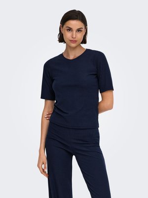 Femme aux cheveux courts et foncés portant un haut côtelé bleu marine à manches courtes et un pantalon assorti sur un fond clair uni.