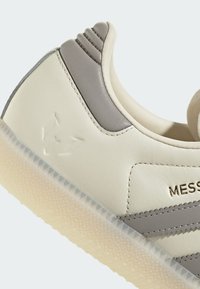 Sneaker de color beige claro con acentos grises, parte superior de cuero liso, tres franjas grises en el costado y suela de goma texturizada. Detalle de logo en relieve.