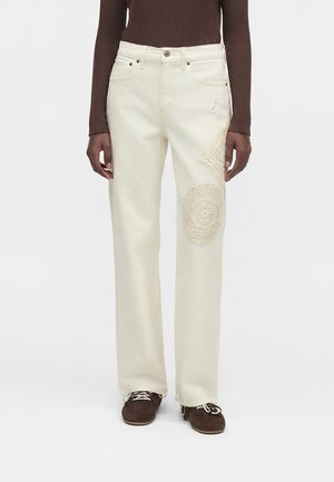 Jean droit taille haute couleur crème avec des détails en patch crocheté sur la cuisse droite, porté avec un haut à manches longues côtelé marron et des chaussures marron.