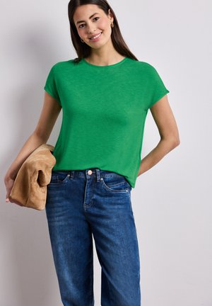 Mujer sonriente, con una camiseta de manga corta verde brillante y jeans azules, sosteniendo una chaqueta marrón doblada en la mano izquierda sobre un fondo liso.