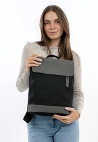 Sac à dos noir avec un rabat gris, fabriqué en matériau durable. Il dispose d'une poignée supérieure et d'accents latéraux. Le design met l'accent sur la simplicité et la fonctionnalité.