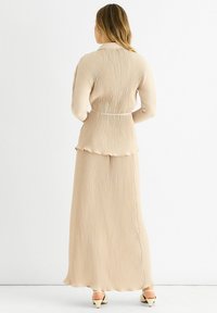Beige, pleated dress with a fitted bodice and flared skirt, featuring long sleeves, a belt at the waist, and a ruffled hemline.