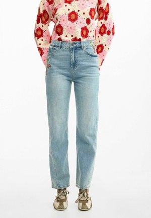 Frau trägt hellblaue High-Waist-Jeans mit floralem Stickmuster, kombiniert mit einem pink-roten Blumen-Crochet-Pullover und braunen Turnschuhen.