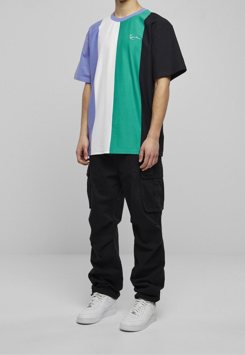 Personne portant une chemise à manches courtes avec des blocs de couleurs lavande, blanc, vert et noir, un pantalon cargo noir et des baskets blanches.