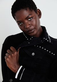 Black denim jacket featuring silver studs along the collar and pockets, with a button closure. White and black striped details on the cuffs.