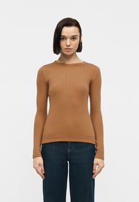 Γυναίκα με κοντά σκούρα μαλλιά που φοράει εφαρμοστή ριγωτή καφέ μακρυμάνικη μπλούζα και σκούρο μπλε τζιν, που στέκεται μπροστά σε απλό λευκό φόντο.