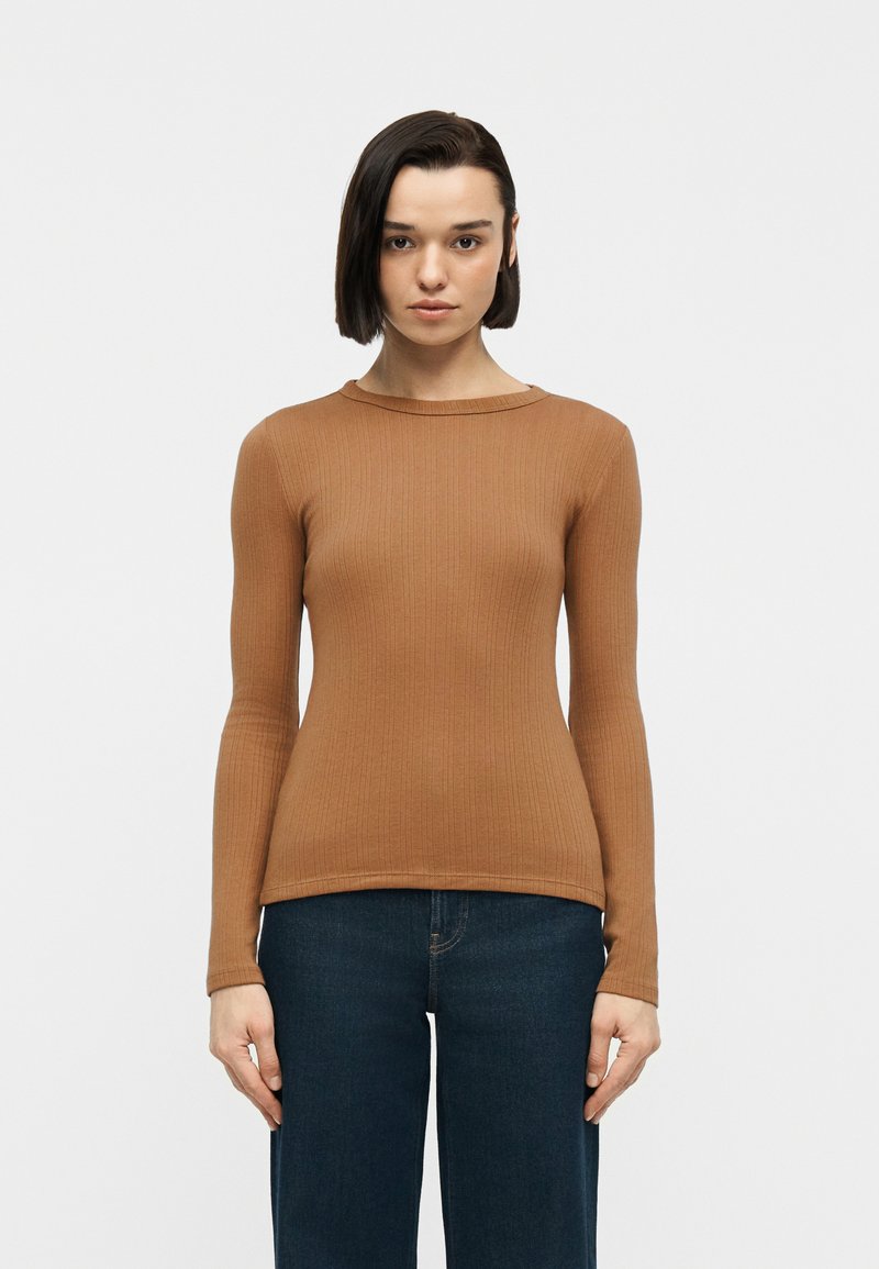 Γυναίκα με κοντά σκούρα μαλλιά που φοράει εφαρμοστή ριγωτή καφέ μακρυμάνικη μπλούζα και σκούρο μπλε τζιν, που στέκεται μπροστά σε απλό λευκό φόντο.