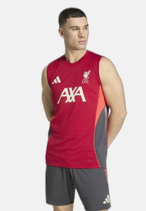 Homme portant un maillot d'entraînement sans manches rouge du Liverpool FC et un short gris foncé, debout les mains derrière le dos, regardant vers la droite.