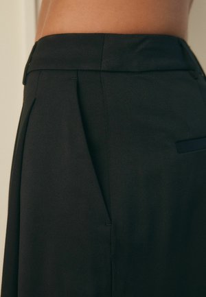 Černé tailored kalhoty s texturovanou tkaninou, plisé vpředu a hladkým pasem. Obsahují boční kapsu s minimálním akcentem.