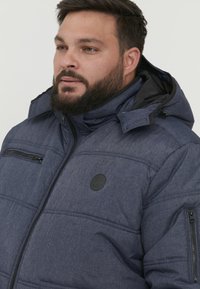 Temno modra puhovka z teksturirano tkanino, z zaklopnimi žepi in kapuco. Ima okrogel logotip na prsih in šivano podrobnost.