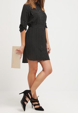 Vestido negro con lunares, cuello con solapa y mangas largas, ceñido en la cintura. Combinado con zapatos de tacón alto negros con tiras y un bolso de mano beige.