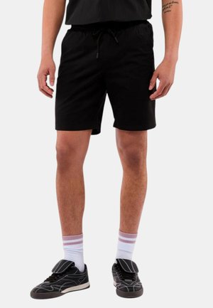 Zwarte shorts met een elastische tailleband en trekkoord, met een gladde textuur en een knielange snit. Draagt zwarte sneakers en witte sokken.