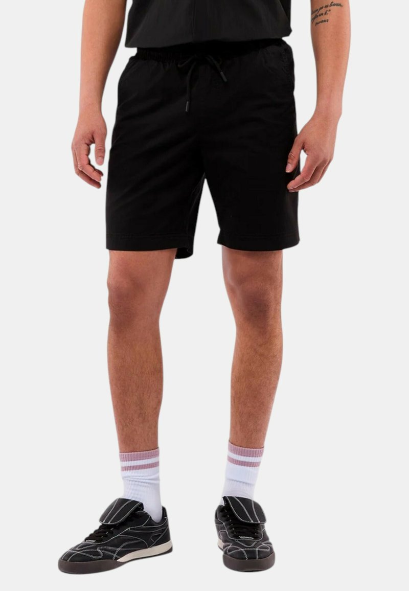 Svarta shorts med elastisk midjeband och dragsko, med en slät yta och knälång design. Bärs med svarta sneakers och vita strumpor.