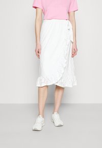 Weißer Wickelrock mit gerafftem Saum, Lochmusterdetails und Bindeschluss, kombiniert mit einem hellrosa T-Shirt und weißen Sneakers.