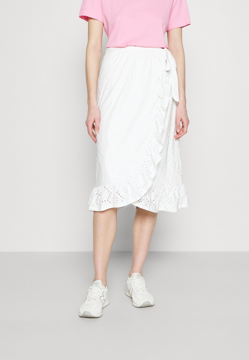 Weißer Wickelrock mit gerafftem Saum, Lochmusterdetails und Bindeschluss, kombiniert mit einem hellrosa T-Shirt und weißen Sneakers.