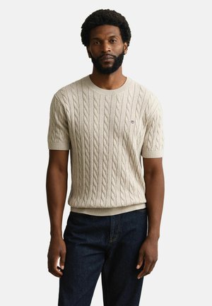 Schwarzer Mann mit Bart, der einen beigen kurzärmeligen Pullover mit Zopfmuster und dunkelblaue Jeans trägt, steht vor einem schlichten hellen Hintergrund.