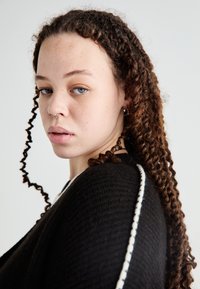 Osoba z kręconymi brązowymi włosami, ubrana w czarny materiałowy element odzieży z białym wykończeniem. Cechy to kolczyk w nosie oraz jasna karnacja.