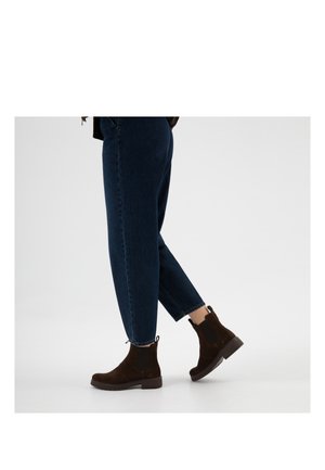 Osoba ubrana w ciemnoniebieskie dżinsy i ciemnobrązowe botki na kostkę, idąca na tle jednolitego jasnego tła.