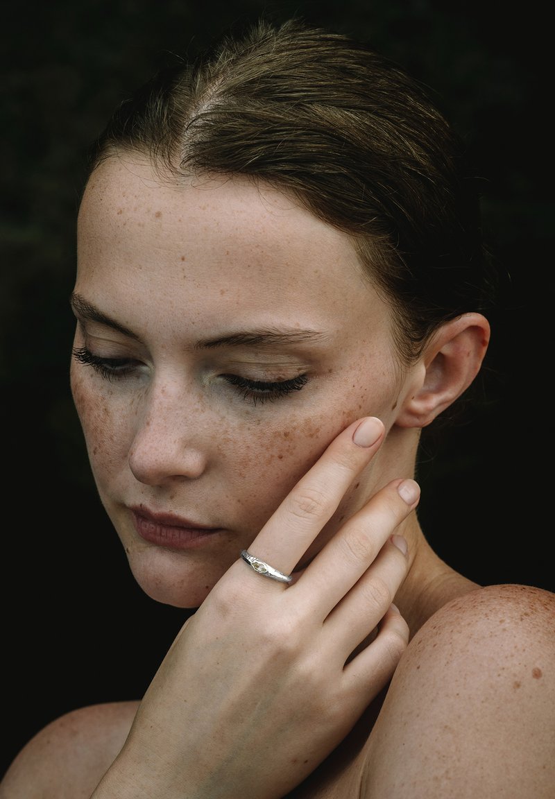 Anillo de plata con un diseño intrincado en un dedo delgado, contra un fondo de piel ligeramente pecosa y tonos oscuros.