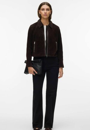 Femme portant une veste en suède marron foncé à fermeture éclair, un pantalon noir, tenant une pochette en cuir noir pliée sur un fond blanc.