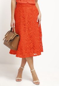 En knä-lång, ljusorange spetskjol med ett blommönster, matchad med en brun handväska och sandaler med remmar.