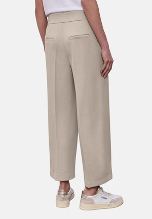 Personne portant un pantalon beige large et court ainsi que des baskets blanches, debout avec les mains détendues le long du corps.
