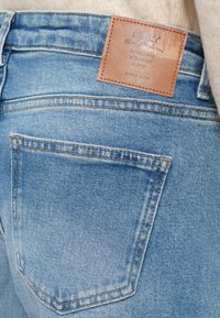 Svijetloplave traperice s smešnim kožnim oznakom na pojasu i stražnjem dzepu. Materijal pokazuje izblijedjeli uzorak s detaljima šivanja.