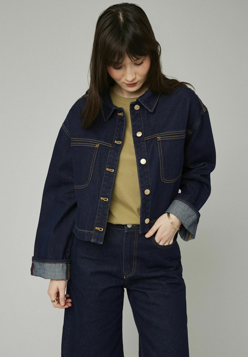 Veste en denim bleu foncé au design court, boutons métalliques et coutures contrastantes. Comprend deux poches avant et un détail de revers plié.