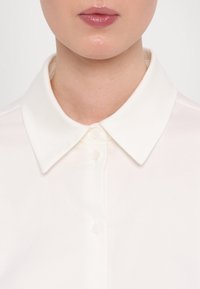 Gros plan d'une personne portant une chemise blanche à col avec les boutons fermés, montrant les lèvres et la partie inférieure du visage sans autres caractéristiques visibles.