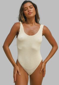 Maillot de bain une pièce couleur crème avec un design texturé en tricot. Présente un décolleté large et des bords festonnés sur les bretelles.