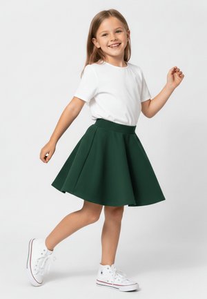 Uśmiechnięta dziewczyna w białej koszulce, ciemnozielonej spódnicy i białych wysokich tenisówkach pozująca z uniesioną jedną nogą na jednolitym tle.