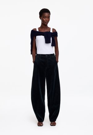 Mujer vistiendo una camiseta sin mangas blanca, suéter azul marino atado alrededor de los hombros, pantalones anchos negros y sandalias negras, de pie con las manos en los bolsillos.