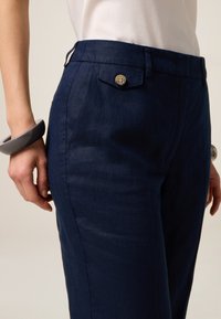 I pantaloni in lino navy presentano una vestibilità slim, chiusura con bottone frontale e tasche laterali. La texture è liscia, con una leggera lucentezza.