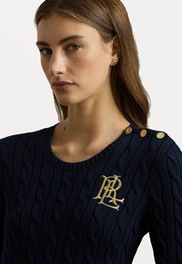 Maglione blu navy con texture a treccia, collo rotondo, ricamo del logo dorato e tre bottoni dorati decorativi sulla spalla.